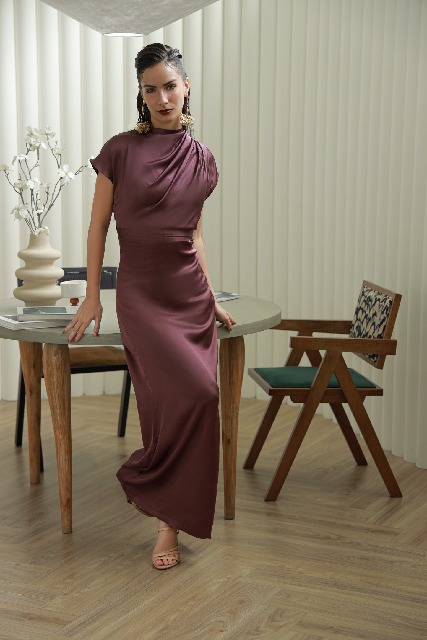 Woman in a long burgundy dress standing in a room with wooden furniture and decor.