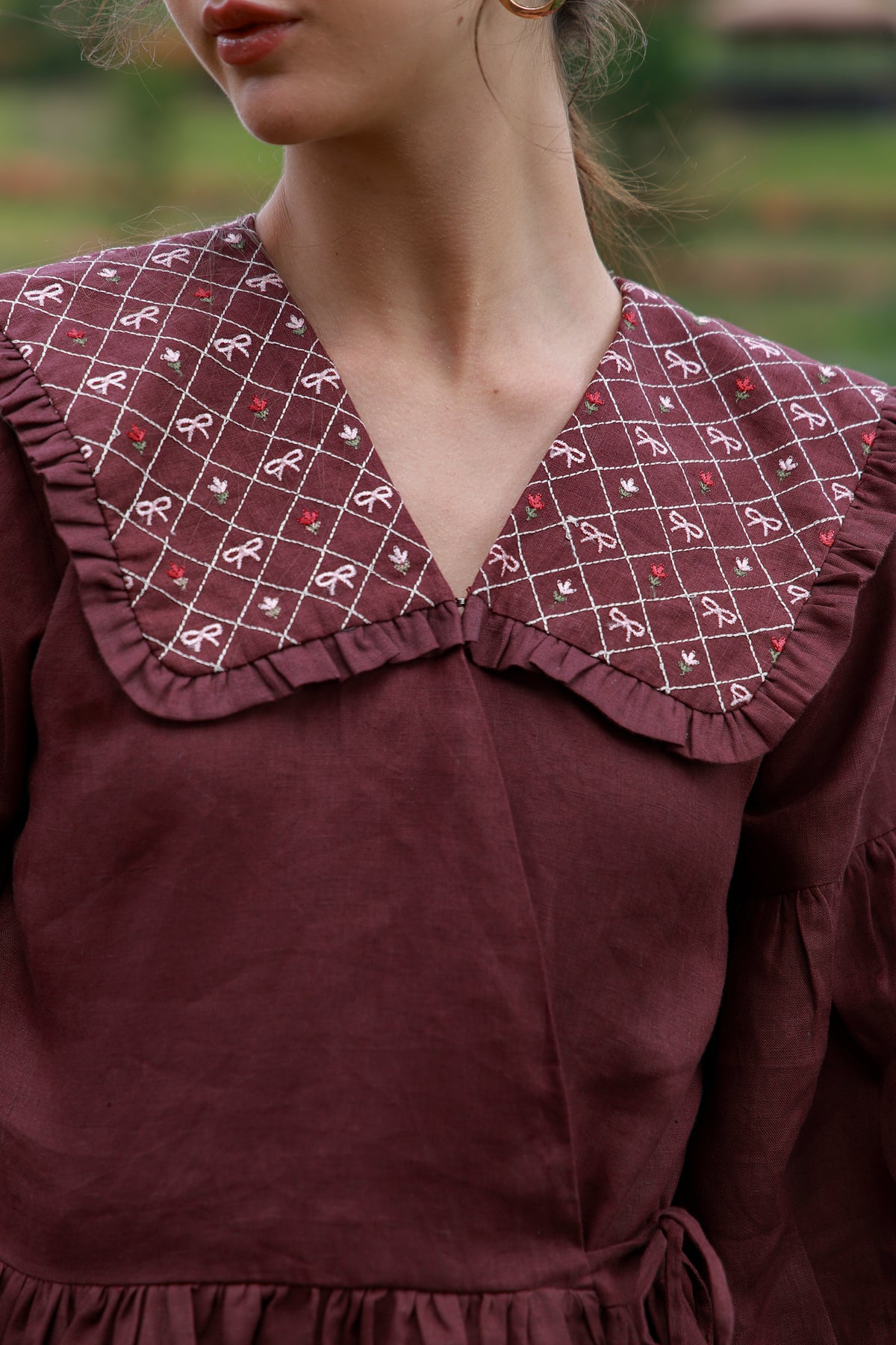 Close-up of a person wearing a burgundy dress with a large ruffled collar featuring a white grid pattern and small red bow motifs, photographed outdoors with a blurred green background.