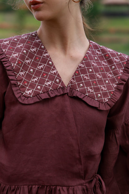 Close-up of a person wearing a burgundy dress with a large ruffled collar featuring a white grid pattern and small red bow motifs, photographed outdoors with a blurred green background.